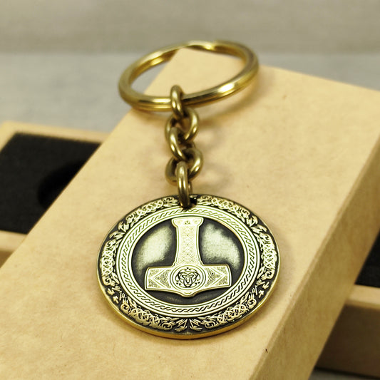 Close-up of a brass Thor's Hammer coin keychain with deep laser engraving, antique patina finish, and decorative border on a craft gift box.