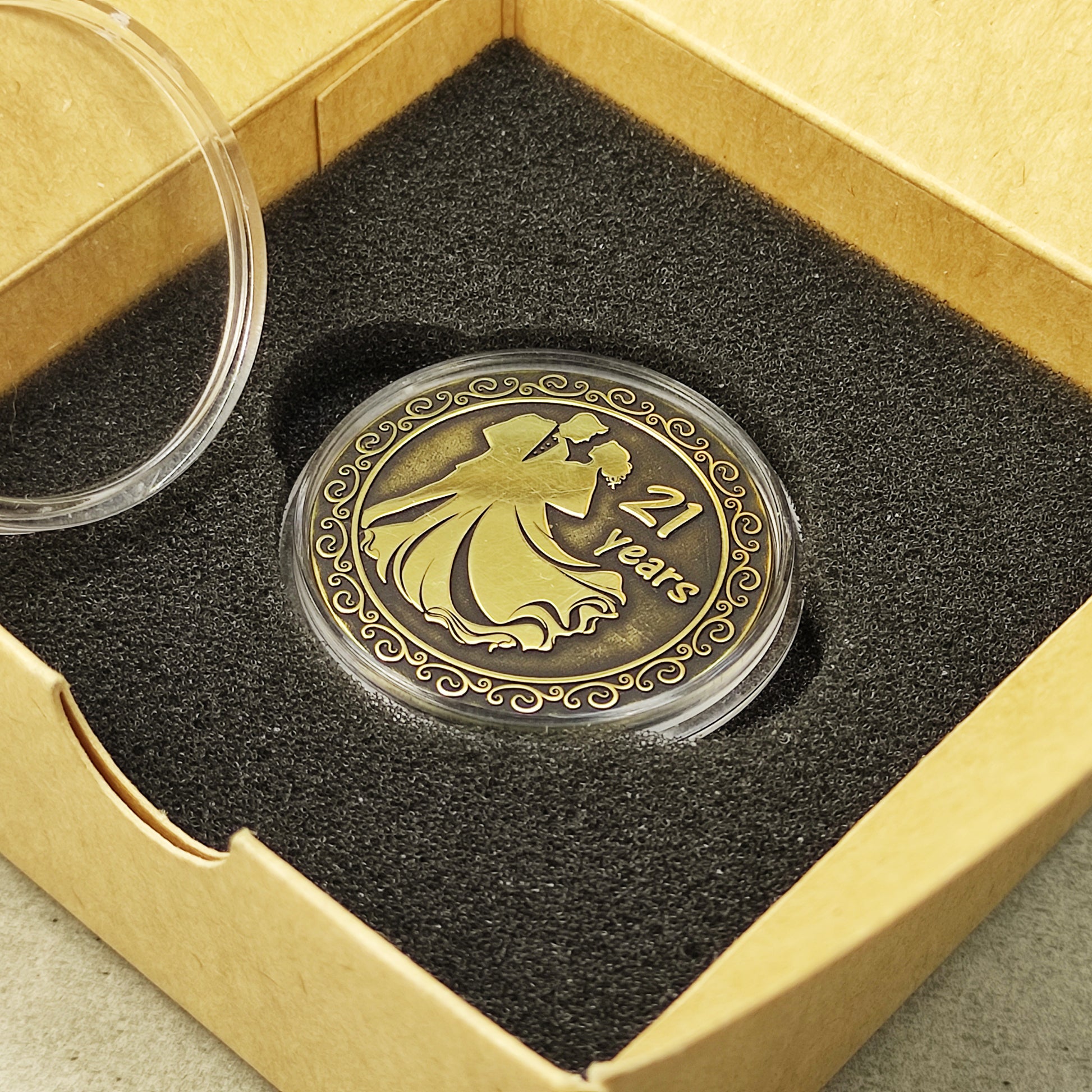 Brass anniversary token with dark patina finish placed in a black foam insert of a square cardboard gift box.