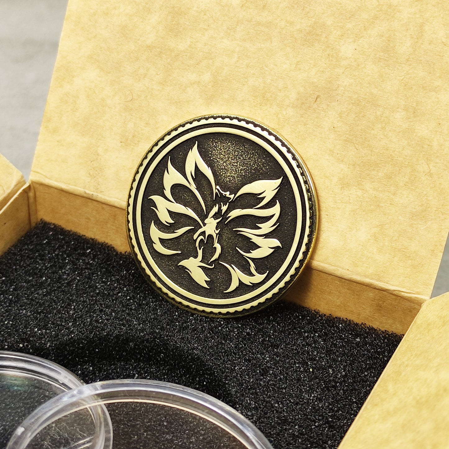Perspective shot of the Kitsune nine-tailed fox coin in brass, leaning against a premium kraft cardboard gift box.